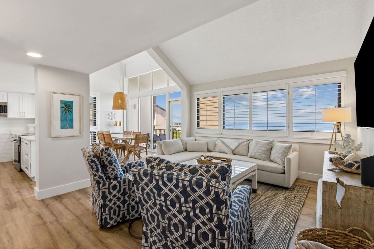 Bright coastal living room with patterned armchairs, white sectional sofa, large windows with ocean view, wooden dining table, and open kitchen. Light wood floors and beach-themed decor create a relaxing, airy atmosphere.