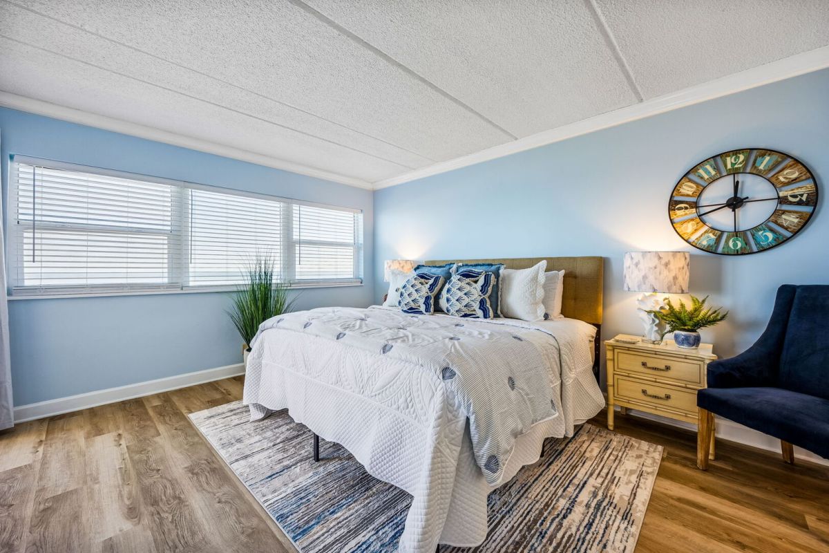 A bright bedroom with light blue walls, a large bed with white and blue bedding, a navy chair, plants, a wooden nightstand, lamps, a clock on the wall, and sunlight streaming through wide windows.