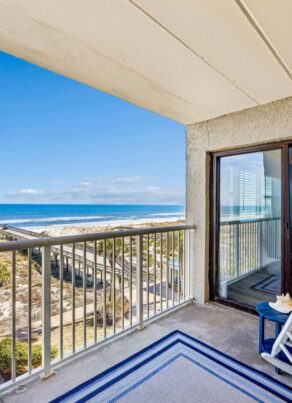 A beachfront balcony with white chairs and a blue rug overlooks the ocean, sandy dunes, and a long wooden pier. A cozy bedroom with a bed and lamp is visible through the sliding glass door.
