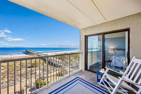 A beachfront balcony with white chairs and a blue rug overlooks the ocean, sandy dunes, and a long wooden pier. A cozy bedroom with a bed and lamp is visible through the sliding glass door.