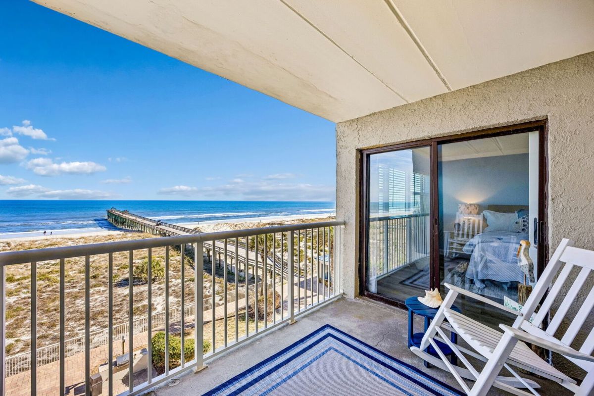 A beachfront balcony with white chairs and a blue rug overlooks the ocean, sandy dunes, and a long wooden pier. A cozy bedroom with a bed and lamp is visible through the sliding glass door.