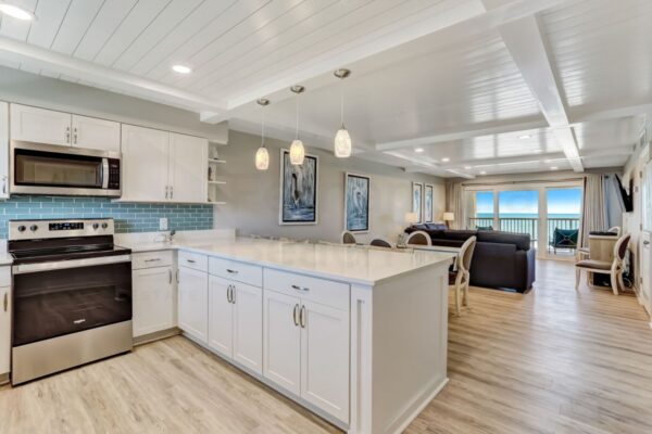 Modern open kitchen and living area with white cabinets, blue tile backsplash, pendant lights, and ocean view through large windows. Light wood flooring and coastal decor create a bright, airy atmosphere.