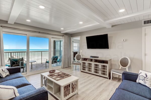 Bright coastal living room with blue sofas, a wall-mounted TV, and large glass doors opening to a balcony with ocean views. Light wood flooring and white paneled ceiling create an airy, inviting atmosphere.