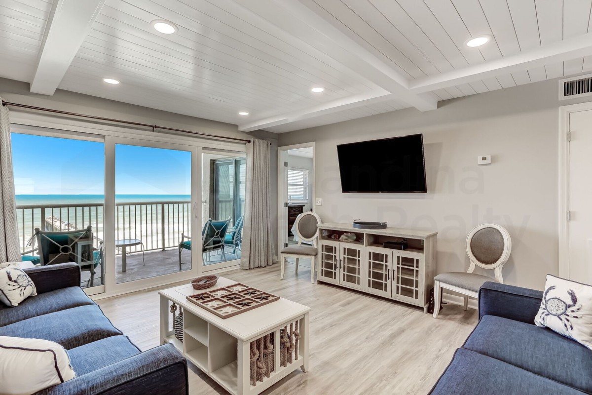 Bright coastal living room with blue sofas, a wall-mounted TV, and large glass doors opening to a balcony with ocean views. Light wood flooring and white paneled ceiling create an airy, inviting atmosphere.