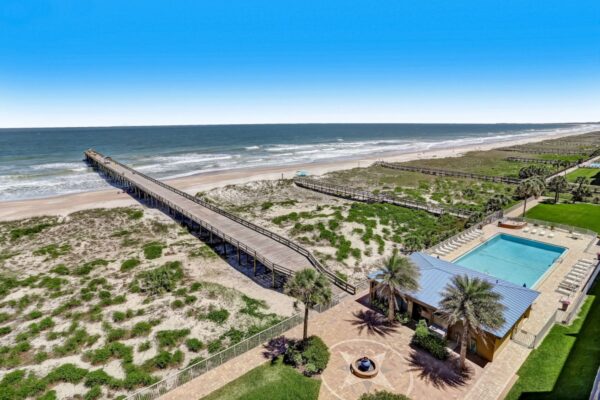 Aerial view of a beach with a long wooden boardwalk, sandy dunes, and a blue swimming pool next to a yellow building with palm trees under a clear blue sky.
