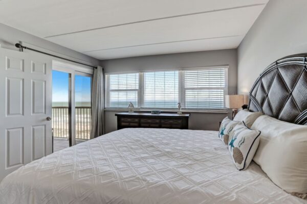A bedroom with a large bed, patterned headboard, pillows, dresser, and windows showing a beach view. A sliding door leads to a balcony with ocean scenery.