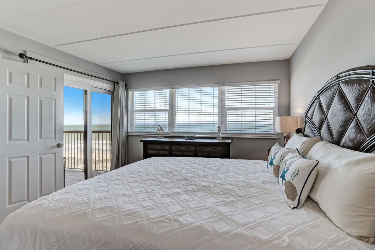 A bedroom with a large bed, patterned headboard, pillows, dresser, and windows showing a beach view. A sliding door leads to a balcony with ocean scenery.