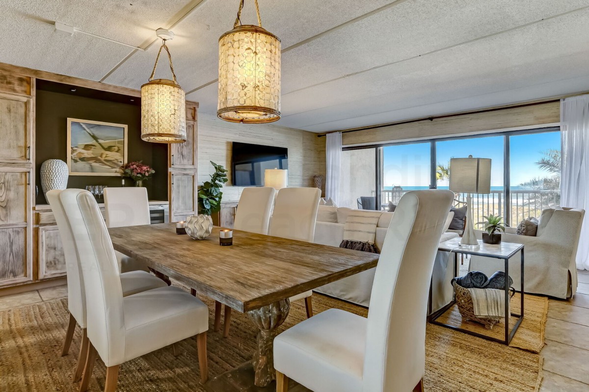A cozy dining and living area with a rustic wooden table, white upholstered chairs, wicker pendant lights, a TV, large windows, and natural decor, overlooking a sunny outdoor view.