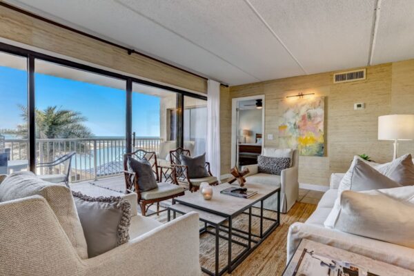 Bright living room with white sofas, armchairs, modern decor, and large windows showing a balcony with an ocean view and palm trees under a clear blue sky.