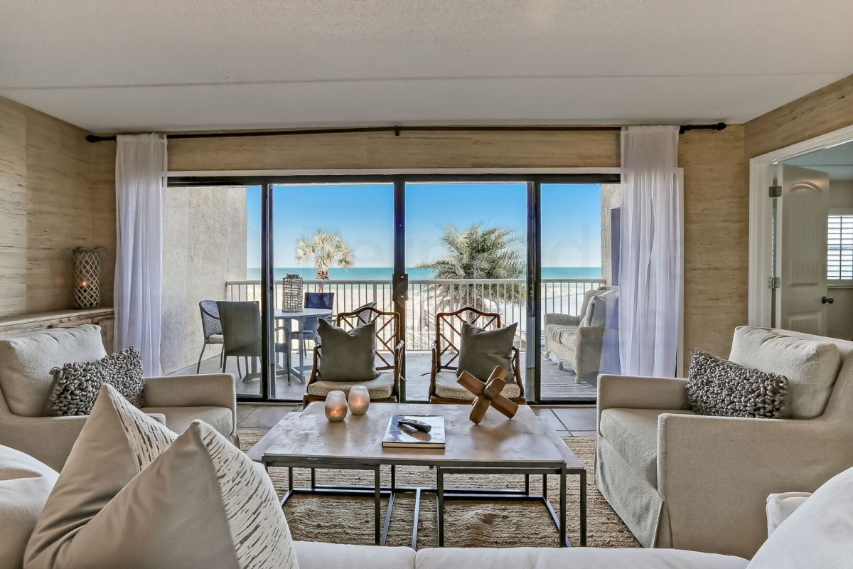 A cozy living room with neutral decor opens to a balcony with chairs, palm trees, and an ocean view under a clear blue sky, seen through large sliding glass doors.