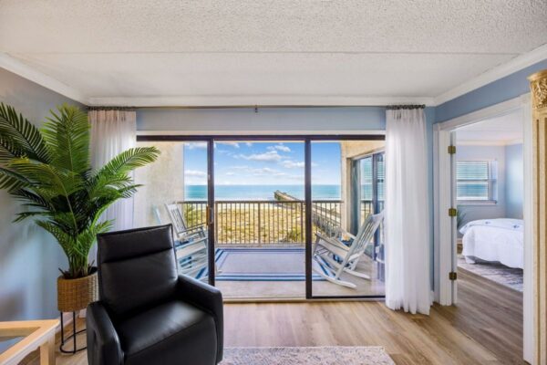 Bright room with a black chair, potted plant, sliding glass doors, and a balcony overlooking the ocean. A bedroom with a white bed is visible through an open doorway on the right.