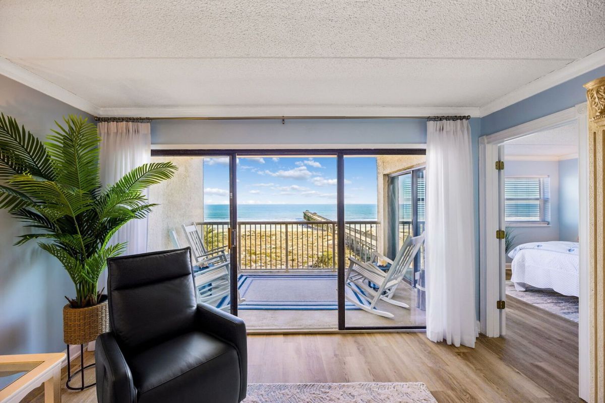 Bright room with a black chair, potted plant, sliding glass doors, and a balcony overlooking the ocean. A bedroom with a white bed is visible through an open doorway on the right.