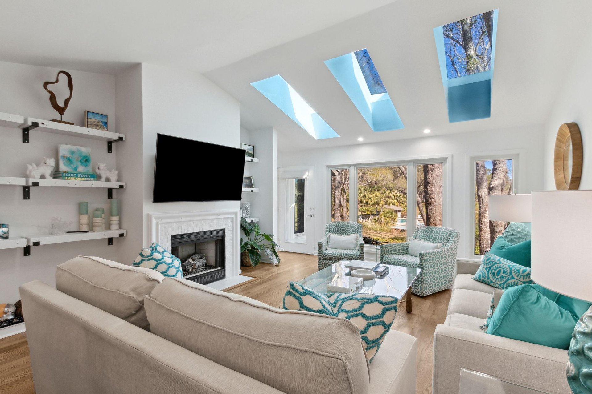 A bright living room with skylights, modern furniture in neutral tones, turquoise accent pillows, patterned armchairs, a wall-mounted TV, fireplace, glass coffee table, and large windows showing trees outside.