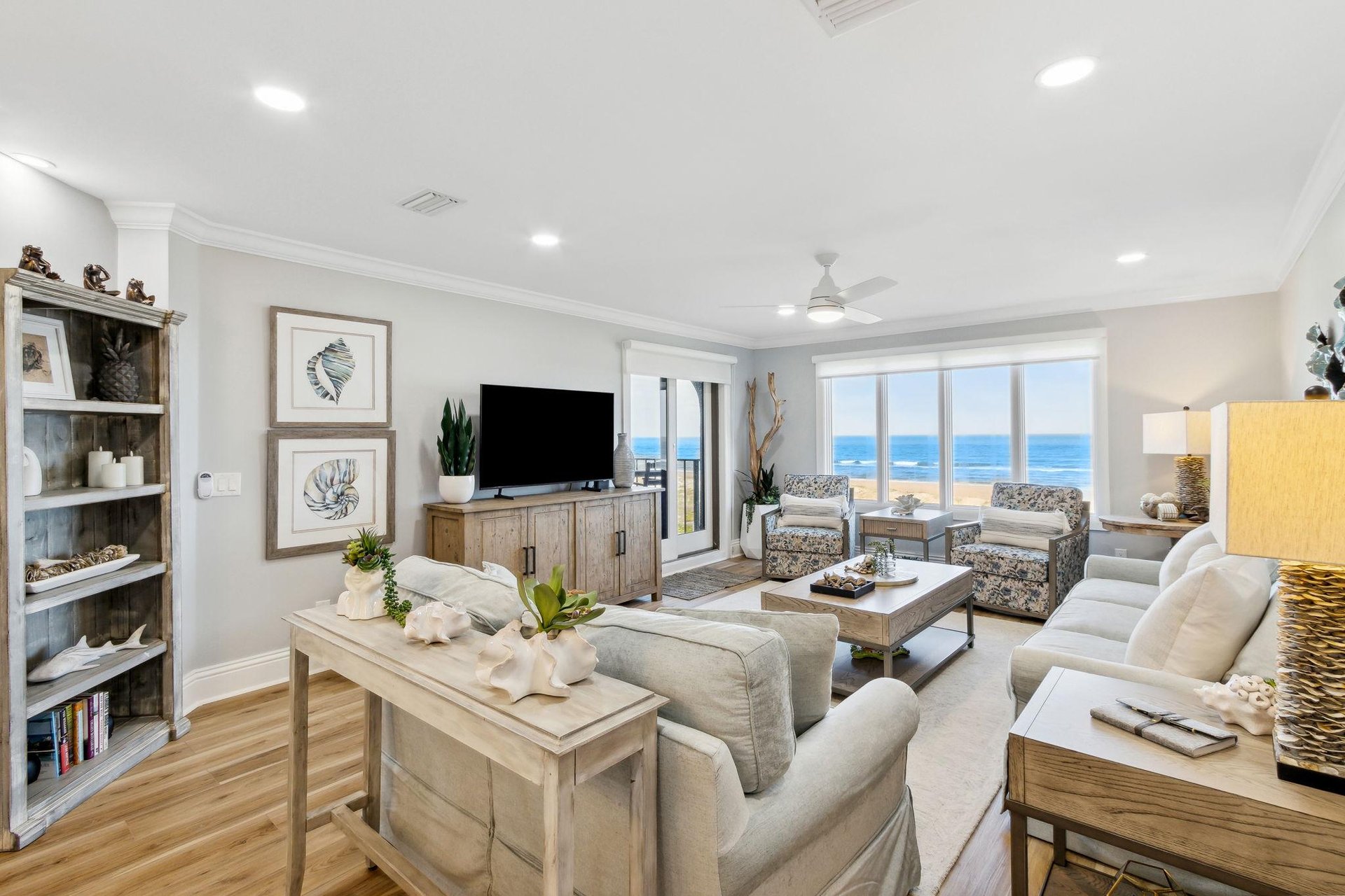 A bright, coastal-themed living room with light furniture, ocean-themed decor, large windows showcasing a beach view, and natural wood accents. There’s a TV, armchairs, and a central coffee table on a light rug.