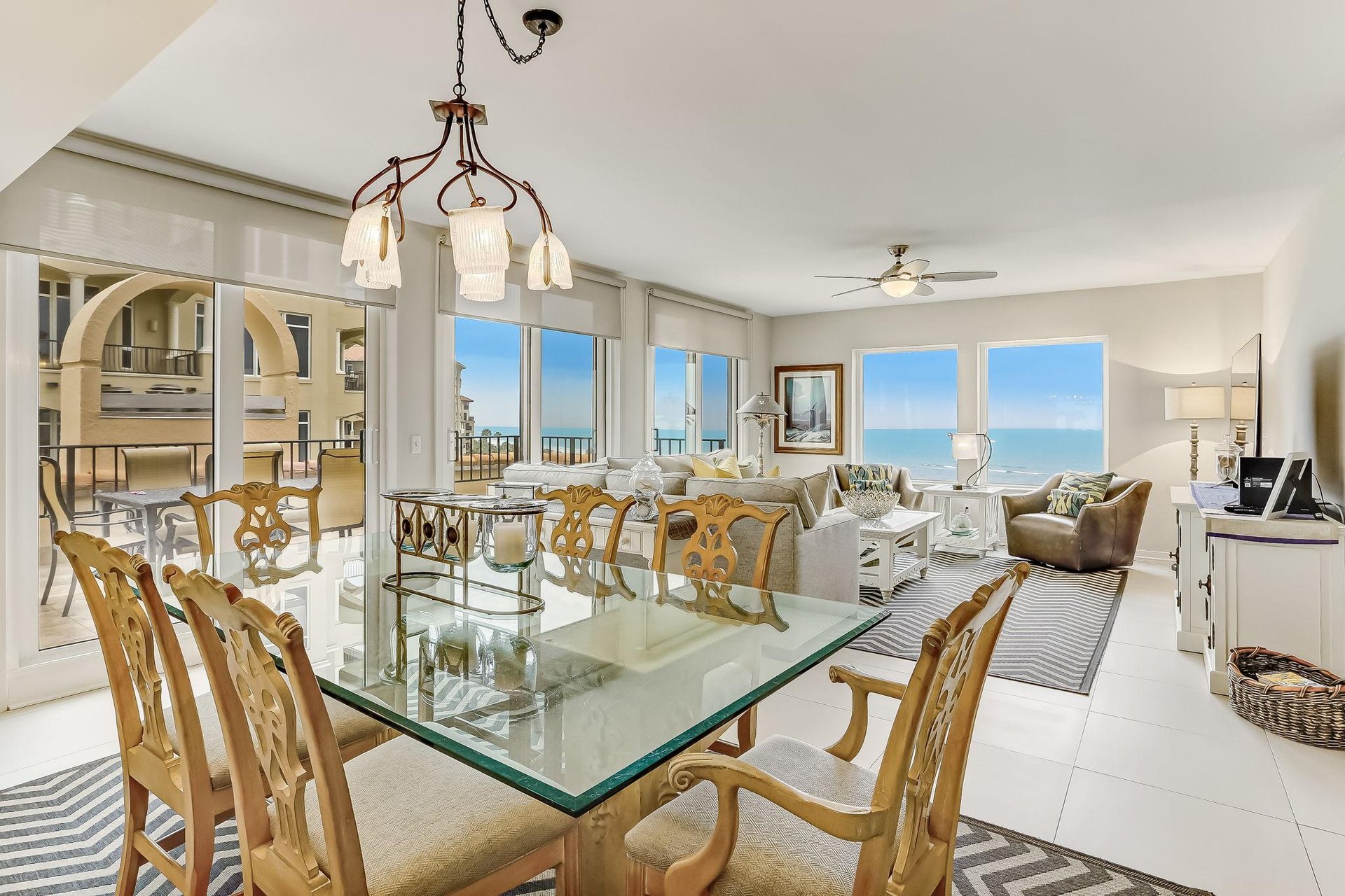 Bright open-plan dining and living area with glass table, wooden chairs, sofas, and large windows overlooking a balcony and ocean view; modern decor with natural light and neutral tones.
