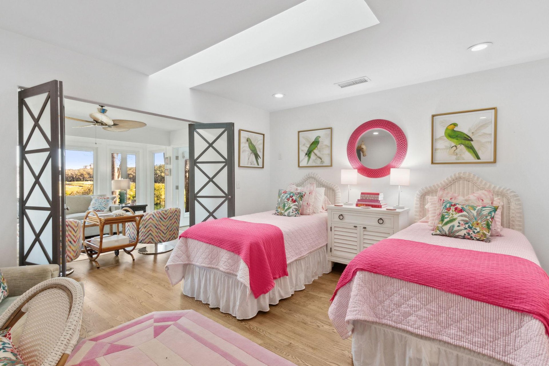 Bright bedroom with two twin beds featuring pink bedding, tropical-themed pillows, and parrot artwork above each bed. A white nightstand with lamps sits between the beds. French doors open to a sunny patio area.