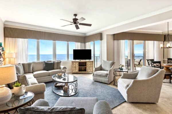 Spacious, modern living room with large windows offering an ocean view, light gray sofas and chairs, a round coffee table, a TV on a wooden stand, and a dining area with glass doors to a balcony.