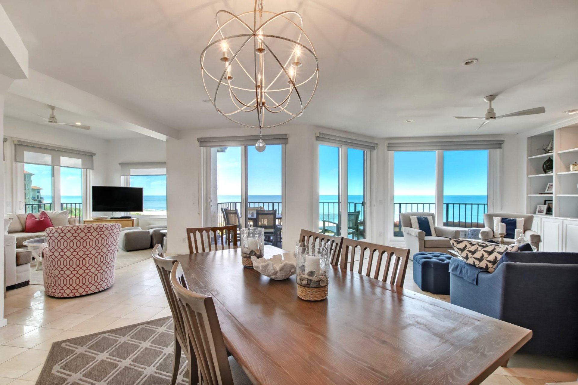 A spacious, modern dining and living area with large windows showcasing an ocean view, wooden dining table with chairs, cozy seating, built-in shelves, and abundant natural light.