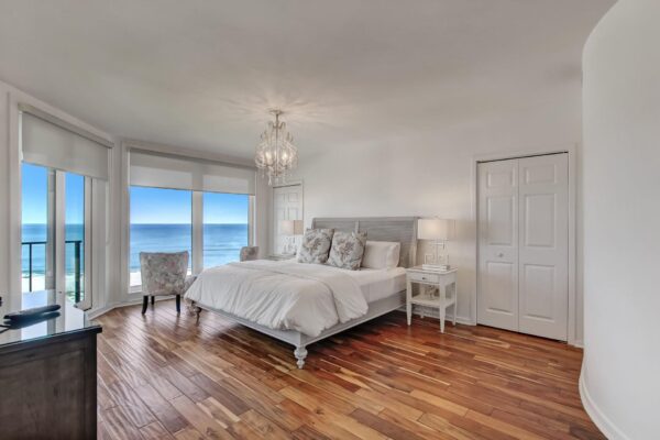 A bright, modern bedroom with wood floors, a white bed, and large windows offering a stunning ocean view. Light walls and a chandelier add elegance, while sunlight fills the room.
