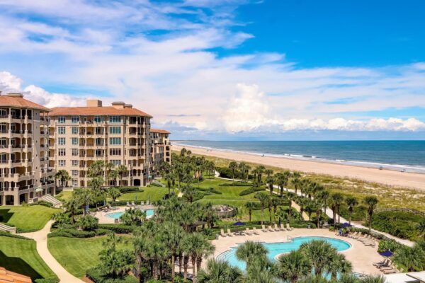 A luxury beachfront resort with palm trees, a large pool, landscaped gardens, and multi-story buildings beside a sandy beach under a bright blue sky with white clouds.