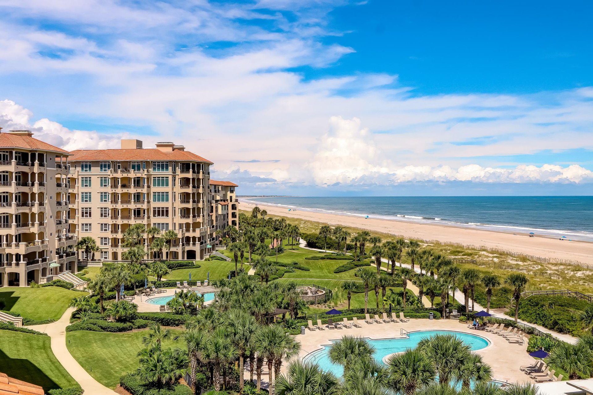 A luxury beachfront resort with palm trees, a large pool, landscaped gardens, and multi-story buildings beside a sandy beach under a bright blue sky with white clouds.