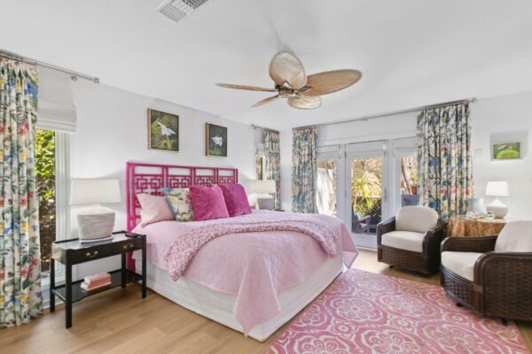 A bright bedroom with a pink bed, floral curtains, pastel decor, and a ceiling fan with leaf-shaped blades. There are two armchairs, large windows, and paintings above the bed, creating a cozy, inviting atmosphere.