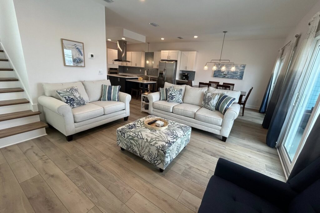 Modern living room with light wood flooring, beige sofas with striped pillows, a patterned ottoman, navy chair, open kitchen, dining area, staircase, and large windows letting in natural light.