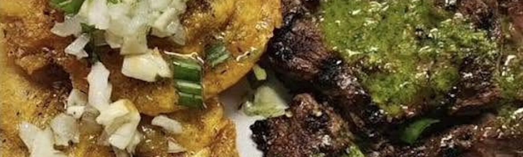 Grilled steak topped with green sauce is served beside two golden fried plantains garnished with chopped onions and herbs on a white plate.