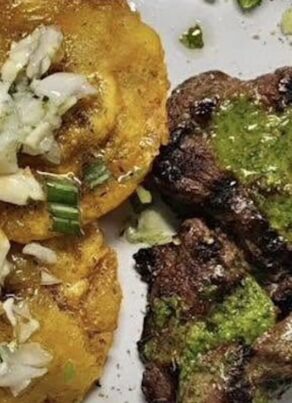 Grilled steak topped with green sauce is served beside two golden fried plantains garnished with chopped onions and herbs on a white plate.
