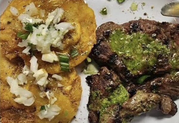 Grilled steak topped with green sauce is served beside two golden fried plantains garnished with chopped onions and herbs on a white plate.