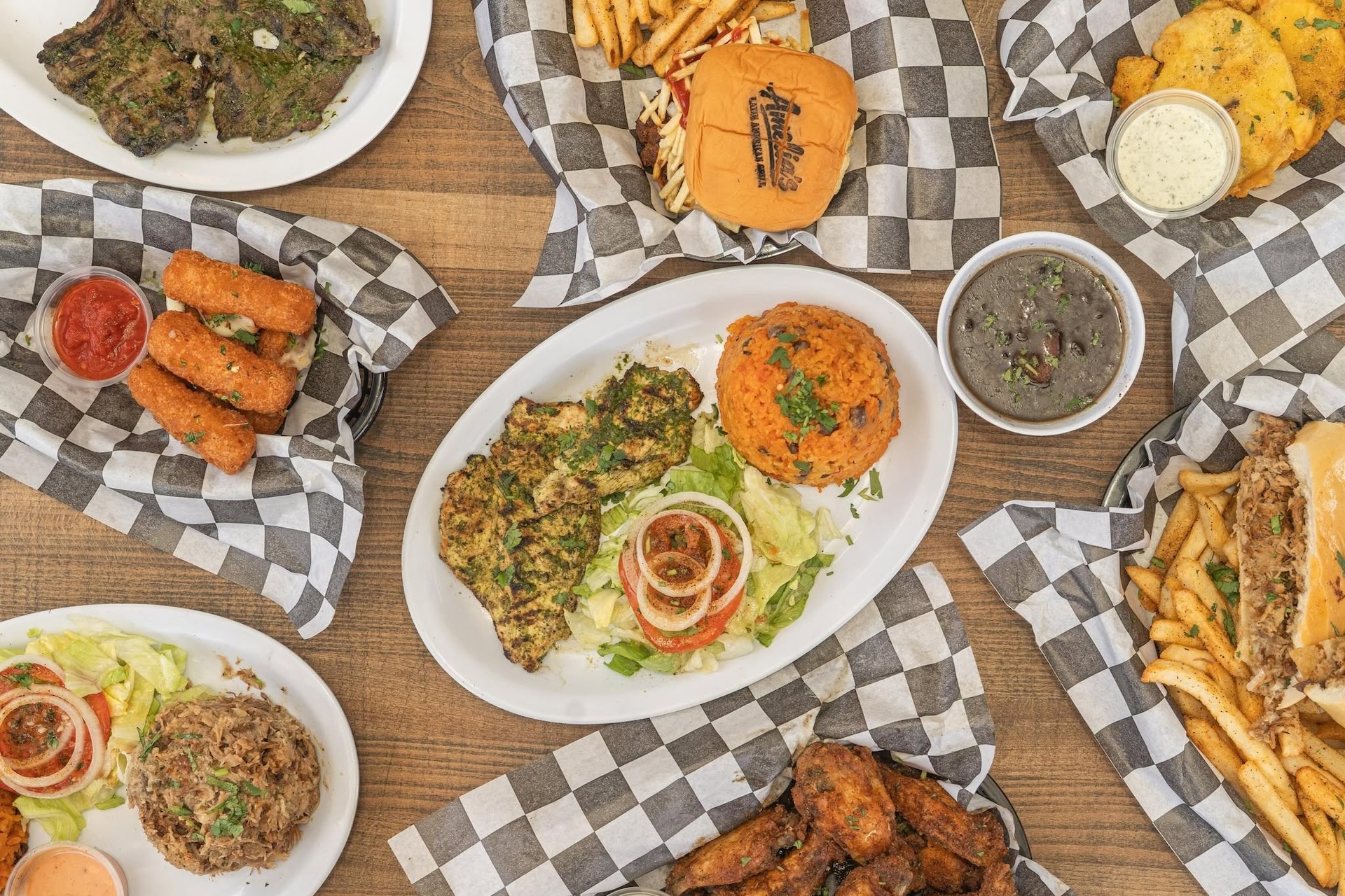 A table with plates of grilled chicken, rice, fries, a burger, fried food, black beans, and wings on checkered paper, showcasing a variety of hearty comfort foods.