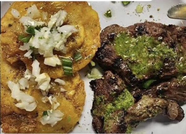 Grilled steak with green sauce sits beside two fried plantains topped with chopped onions and herbs on a white plate.