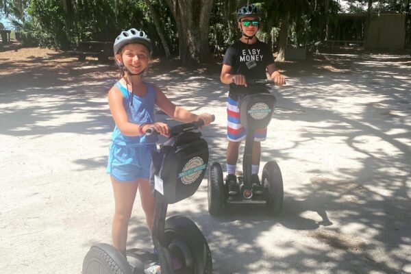 Two children wearing helmets ride Segways on a sunny path surrounded by trees. One wears a blue tank top and shorts; the other wears sunglasses, a black shirt, and striped shorts. Both are smiling and enjoying the outdoors.