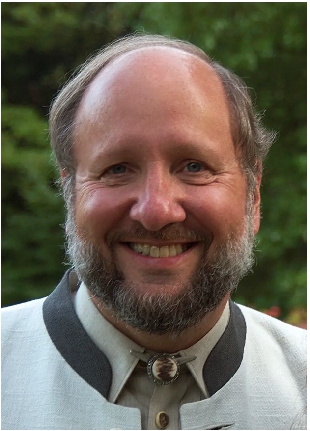 A smiling middle-aged man with a beard and receding hairline wearing a light-colored traditional jacket with a dark trim, standing outdoors with greenery in the background.