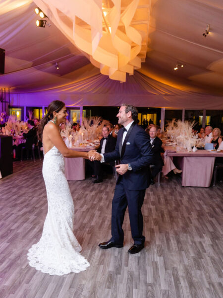A bride in a white lace gown and a groom in a dark suit dance together on a wooden floor at a wedding reception, with guests seated at decorated tables in the background.