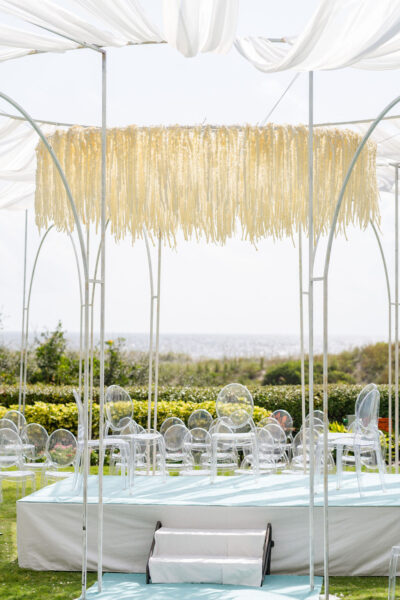 A wedding ceremony setup outdoors features clear acrylic chairs on a light blue platform, under a canopy of white fabric with a hanging fringe decoration, and greenery with ocean visible in the background.