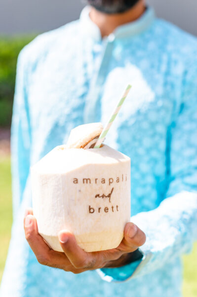 A person in a light blue patterned outfit holds a coconut with a straw. The coconut is engraved with the names amrapali and brett. The background is softly blurred.