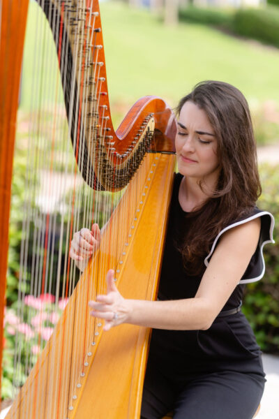 A woman with long brown hair, wearing a black outfit, is playing a large wooden harp outdoors. She appears focused, with a background of greenery and pink flowers.