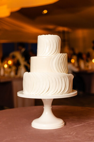 Three-tiered white wedding cake on a white stand, decorated with overlapping circular white fondant accents, set against a softly lit indoor background with candles and blurred guests.