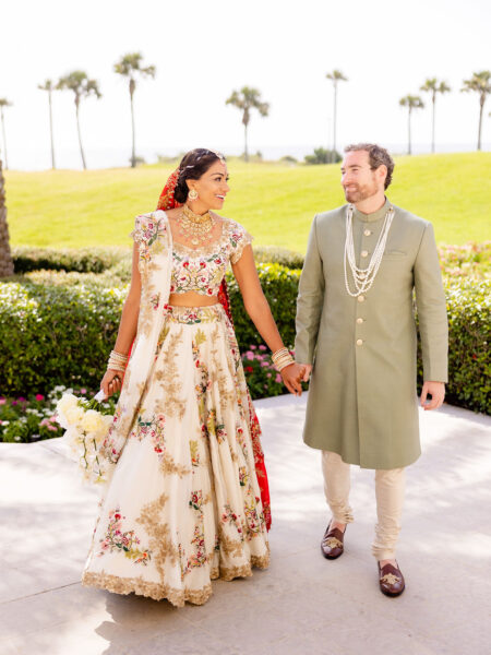 A bride and groom in traditional wedding attire hold hands and smile at each other outdoors, with green grass, palm trees, and the ocean in the background. The bride wears a floral lehenga, and the groom wears a sage green sherwani.