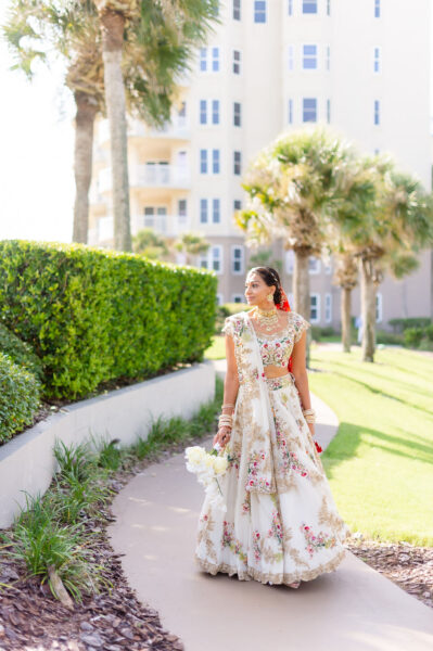 A woman in a white and floral embroidered traditional Indian lehenga, holding a bouquet, walks along a sunlit path with palm trees and a tall building in the background.
