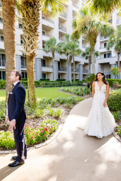 A bride in a white gown stands on a garden path, looking at a groom in a dark suit who faces away from her, surrounded by palm trees and a modern building in the background.