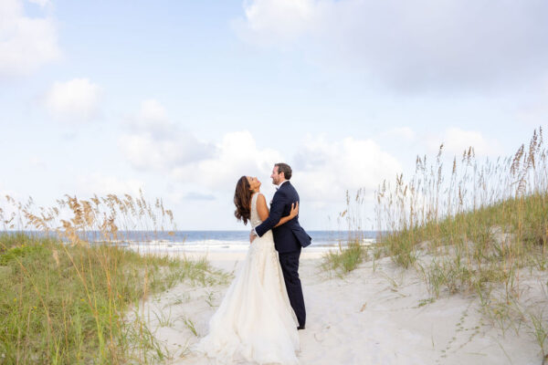 A bride and groom embrace and smile at each other on a sandy beach path, with grass on either side and the ocean visible in the background under a partly cloudy sky.