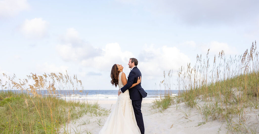 A bride and groom stand on a sandy beach path surrounded by grass, embracing and looking at each other, with the ocean and a blue sky with clouds in the background.
