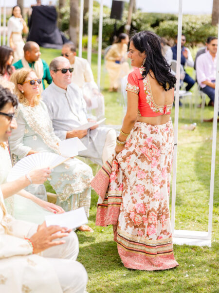 A woman in a floral saree and red blouse stands outdoors, smiling and looking to the side, as seated guests in light-colored traditional attire watch her during a sunny celebration on green grass.
