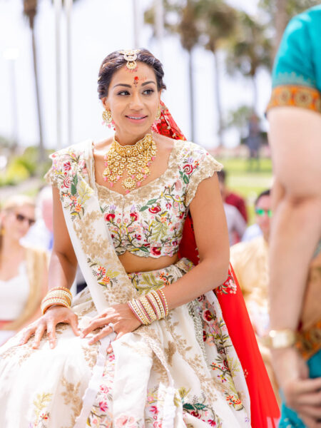 A woman dressed in an ornate cream and red traditional Indian bridal outfit with gold jewelry and floral embroidery sits outdoors, smiling, with blurred palm trees and people in the background.