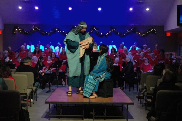 Two people in biblical costumes, one holding a baby, stand on stage in front of a seated choir dressed in red; the stage is decorated with string lights, and the audience is seated facing the performers.
