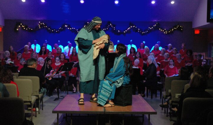Two people in biblical costumes, one holding a baby, stand on stage in front of a seated choir dressed in red; the stage is decorated with string lights, and the audience is seated facing the performers.