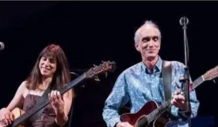 A woman and a man play guitars on stage, both smiling. The woman has long brown hair and wears a sleeveless top, while the man has short gray hair and wears a blue patterned shirt. Microphones are in front of them.