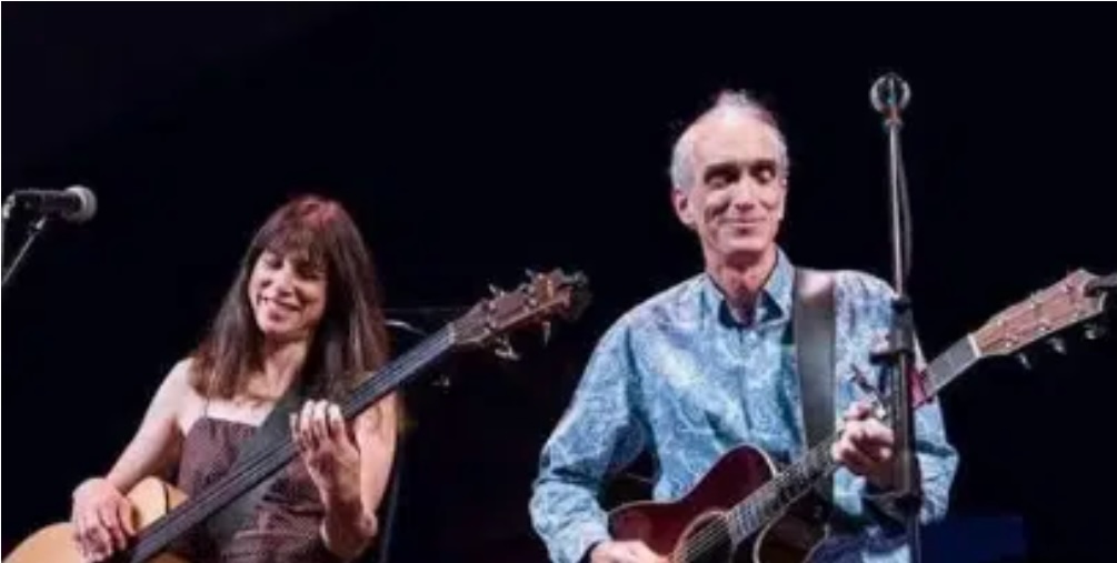 A woman and a man play guitars on stage, both smiling. The woman has long brown hair and wears a sleeveless top, while the man has short gray hair and wears a blue patterned shirt. Microphones are in front of them.